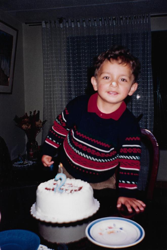 Daniel Felipe Montenegro at two years old, in a striped sweater, standing over a white cake with a lit number-two candle