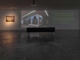 Exhibition view of Visiones inestables: a projected AI-generated image of the museum, a projected quote and a lit photograph in a dark gallery