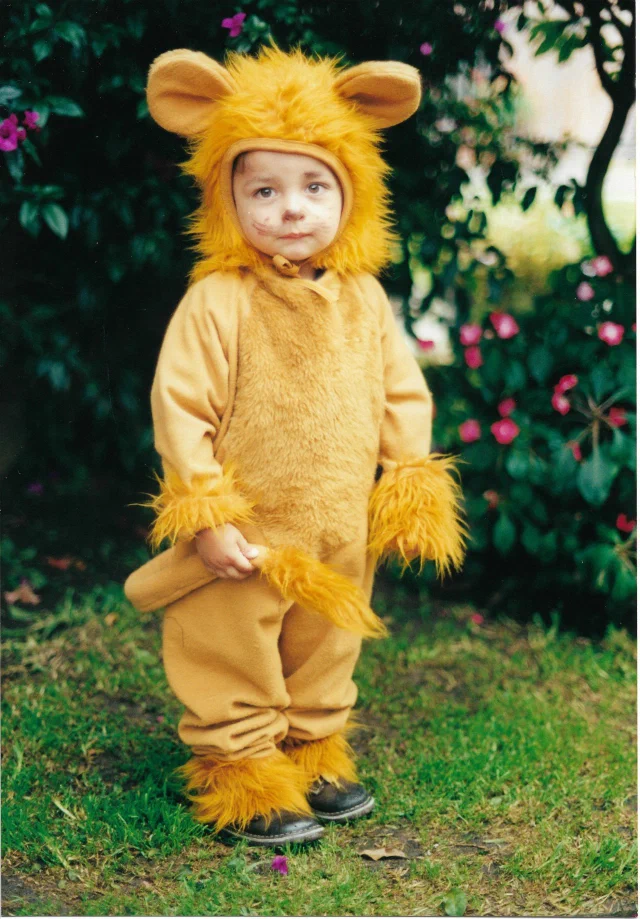 Daniel Felipe Montenegro at three years old, dressed as a lion in a maned hood and furry cuffs, standing on the grass in a garden