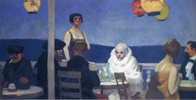 A clown in white make-up sitting alone at a café terrace at night, among other figures under paper lanterns